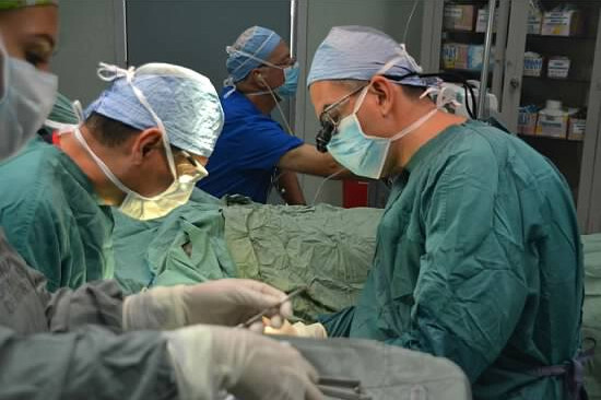 Two physicians are bend over a patient in an operating room. They are wearing surgical scrubs and face masks. Other medical providers can be seen in the background and foreground.